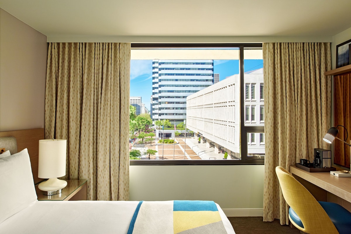 A hotel room with a bed, a desk, a lamp, and a view of buildings through a window.