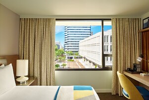 Una camera d'albergo con un letto, una scrivania, una lampada e vista sugli edifici attraverso una finestra.