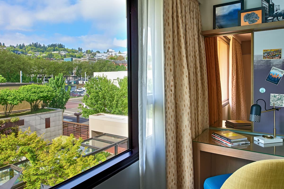 A room with a large window offering a view of a cityscape, a desk with books, a lamp, and a curtain.