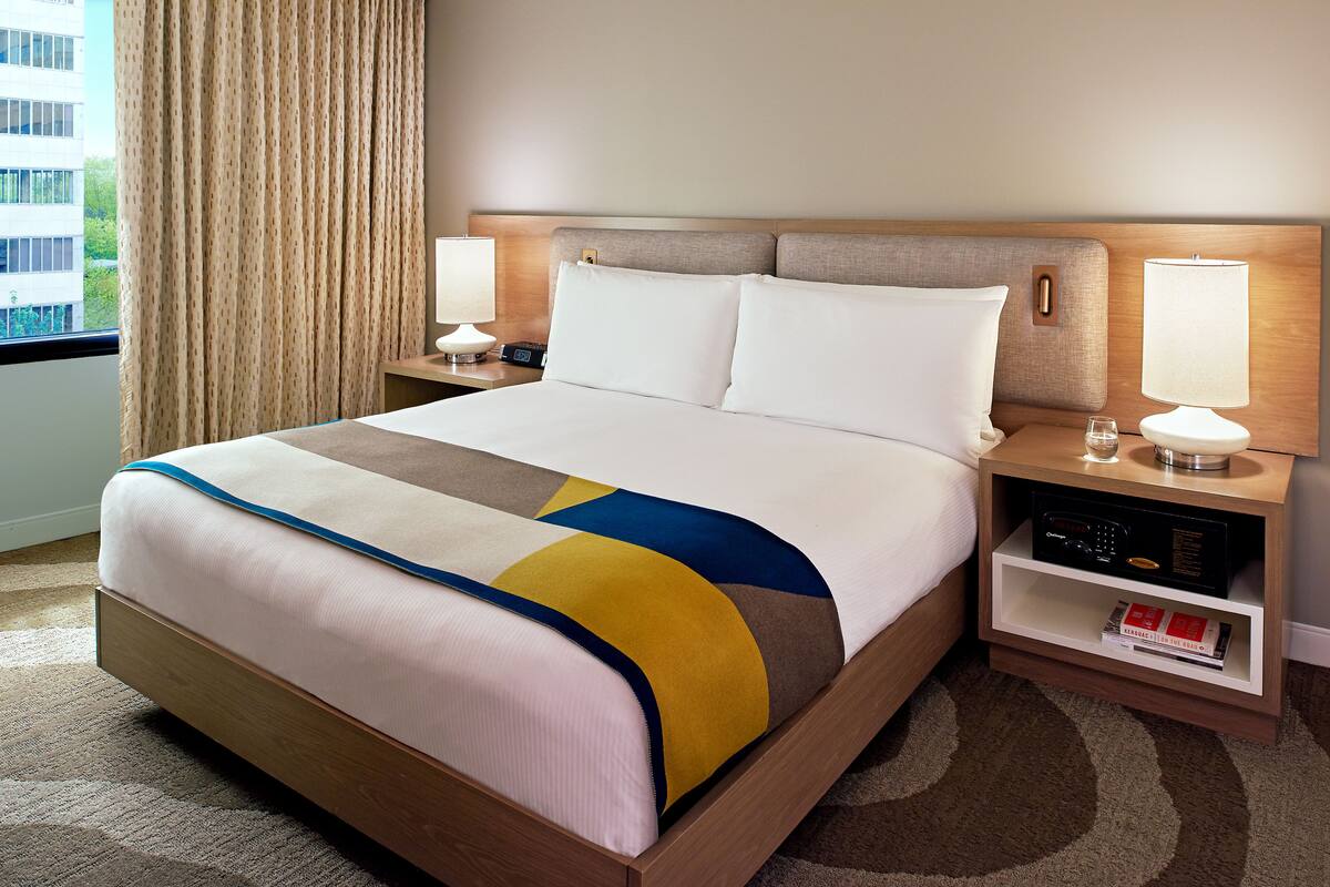 A hotel room with a large bed, two bedside tables with lamps, a bookshelf, and a view of a building outside.