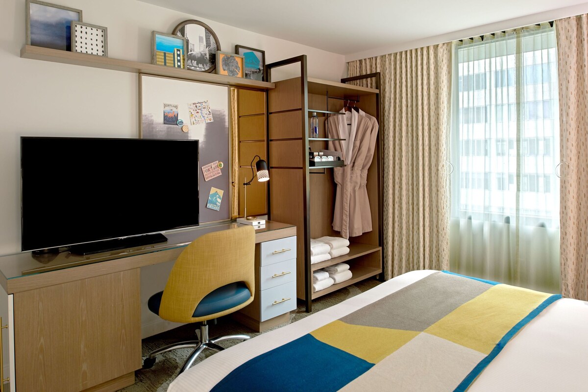A hotel room with a bed, a desk with a TV, a chair, a wardrobe, and a window with curtains.