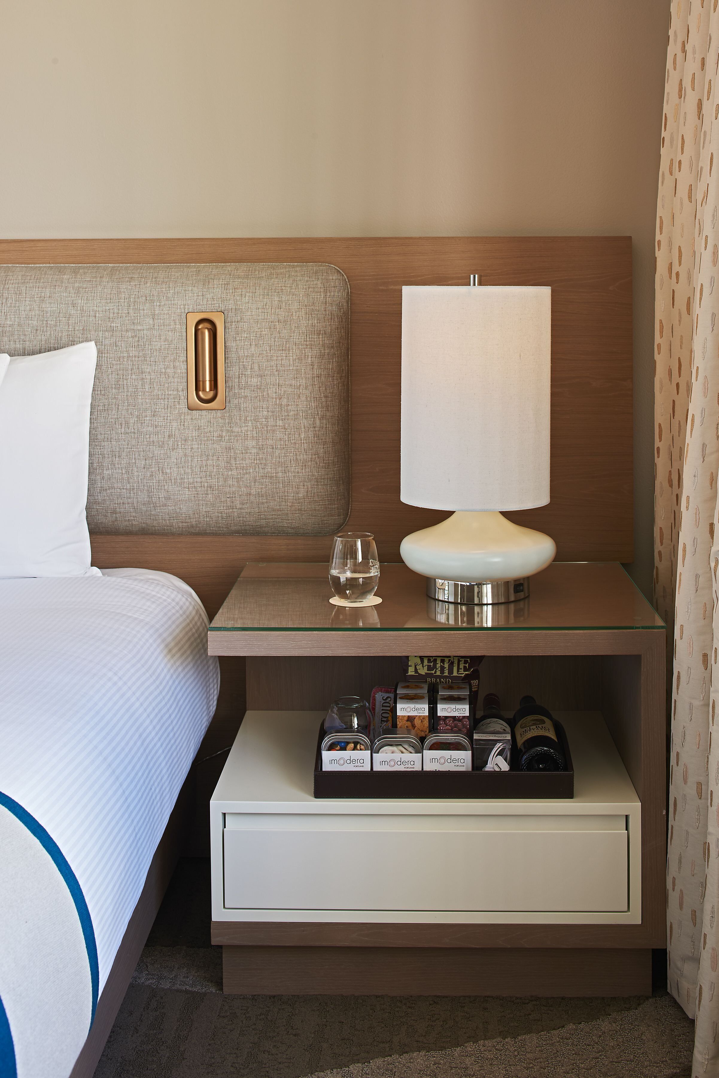 Habitación de hotel ordenada con cama, mesita de noche con lámpara, vaso de agua y minibar.
