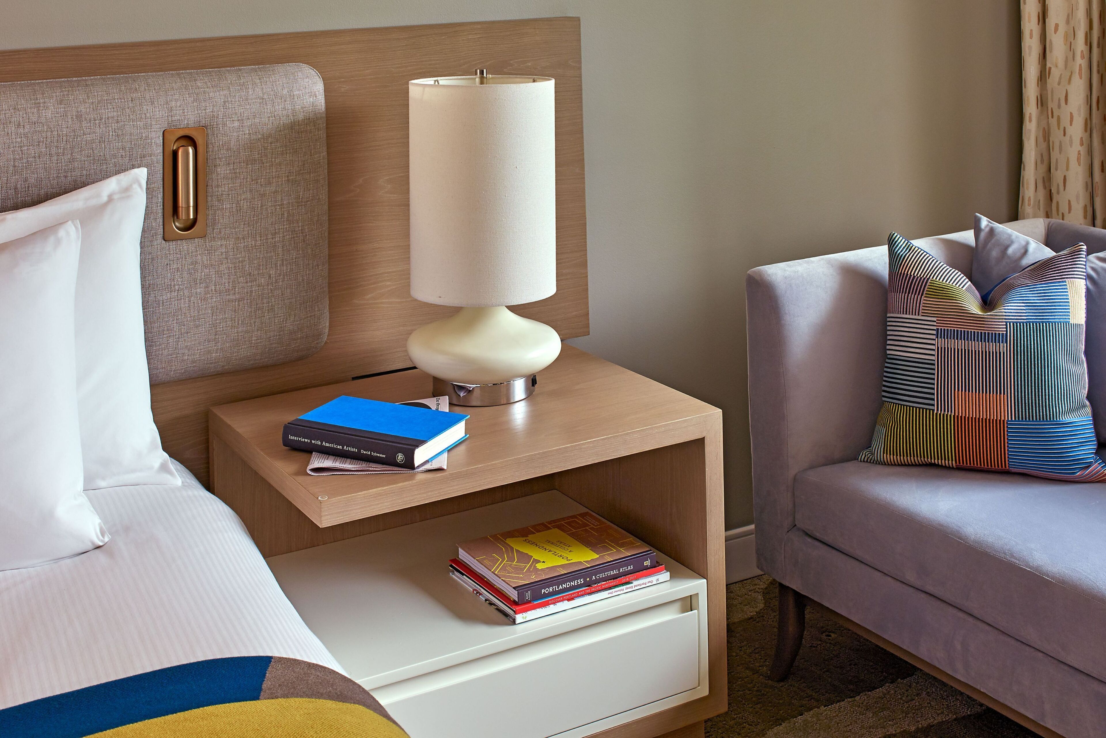 Une chambre d’hôtel bien rangée, avec un lit, une table de chevet dotée d’une lampe, une chaise agrémentée de coussins à motifs et une petite table de nuit avec des livres.