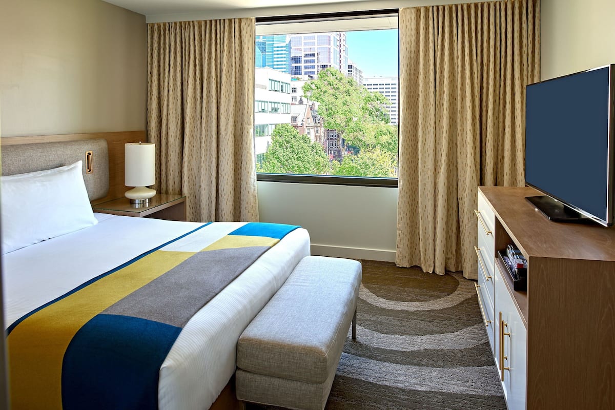 A hotel room with a large bed, a bedside table with a lamp, a TV on a wooden stand, and a window with curtains.