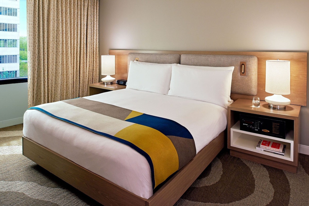 A hotel room with a large bed, two bedside tables with lamps, a bookshelf, and a view of the outside.