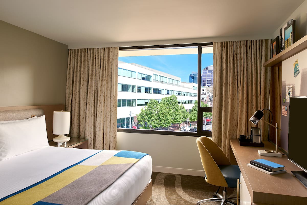 A hotel room with a bed, a desk, a chair, a lamp, and a view of a cityscape through large windows.
