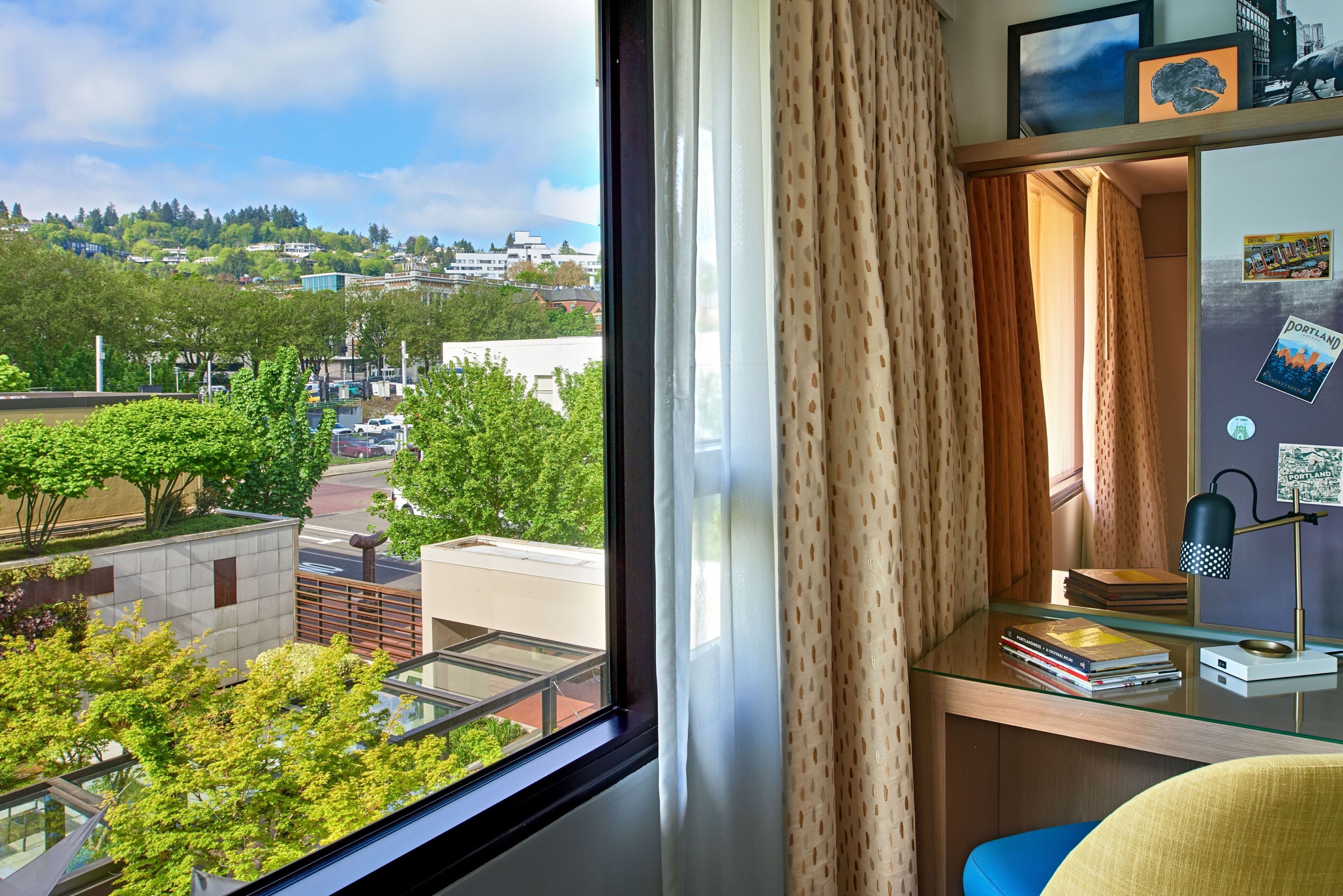 A room with a large window, a desk with books, a lamp, and a view of a cityscape.