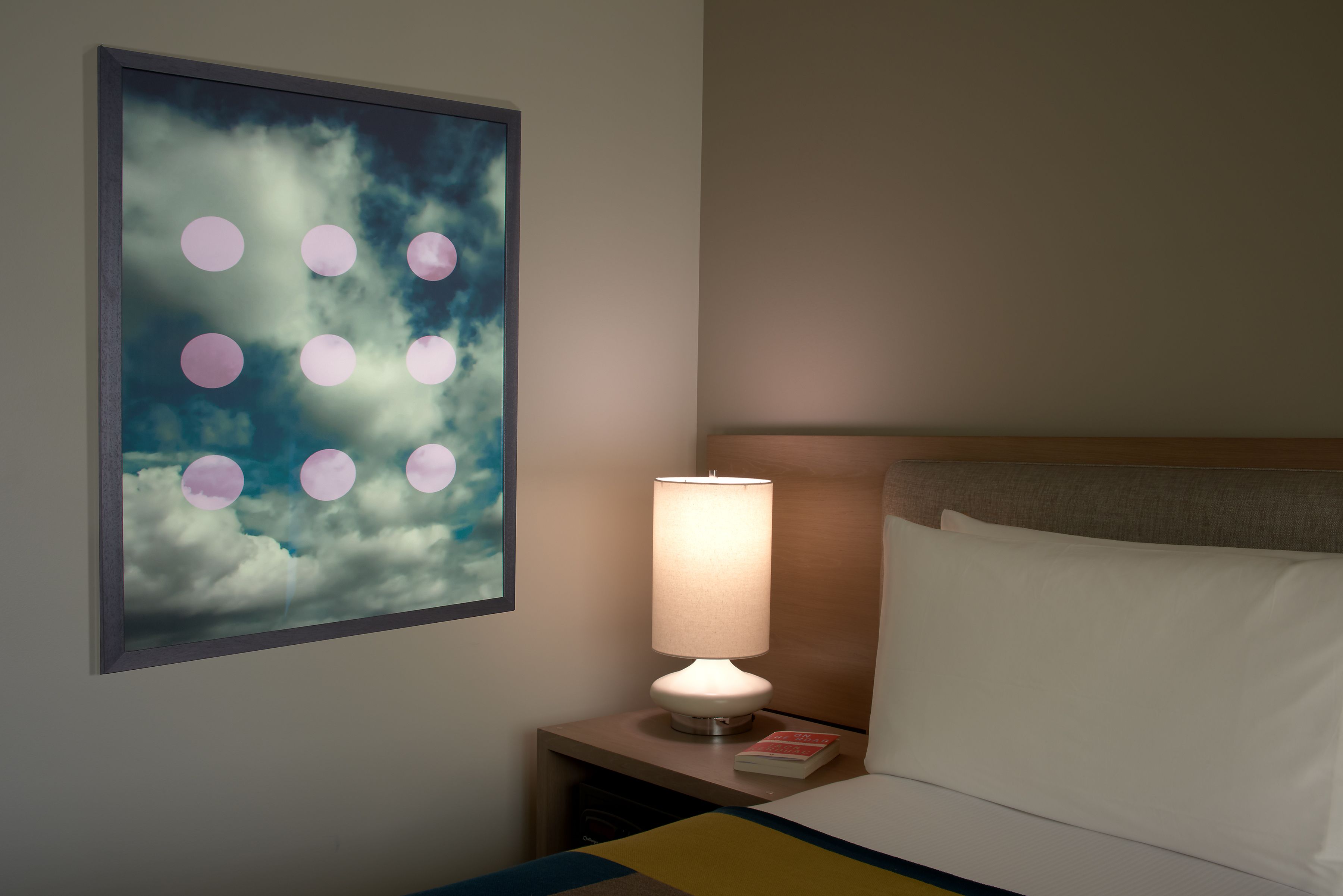 A hotel room with a bed, a nightstand with a lamp, a book, and a wall art of clouds and circles.