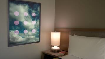 A hotel room with a bed, a nightstand with a lamp, a book, and a wall art of clouds and circles.