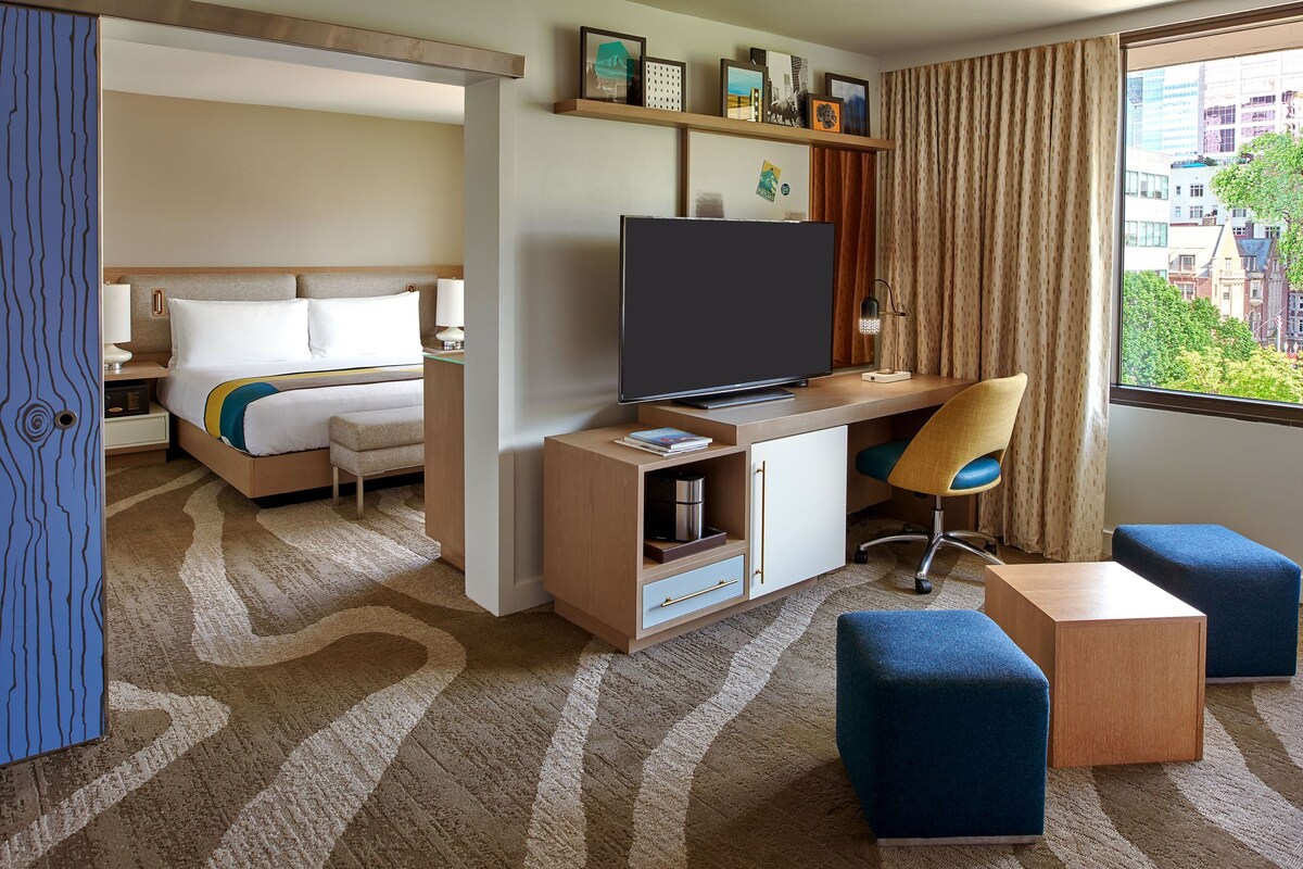 A hotel room with a bed, a desk with a TV, a chair, and a window with a city view.