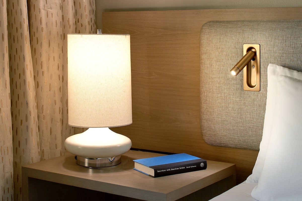 A neatly made hotel room with a bed, a bedside table with a lamp, a book, and a wall-mounted light fixture.