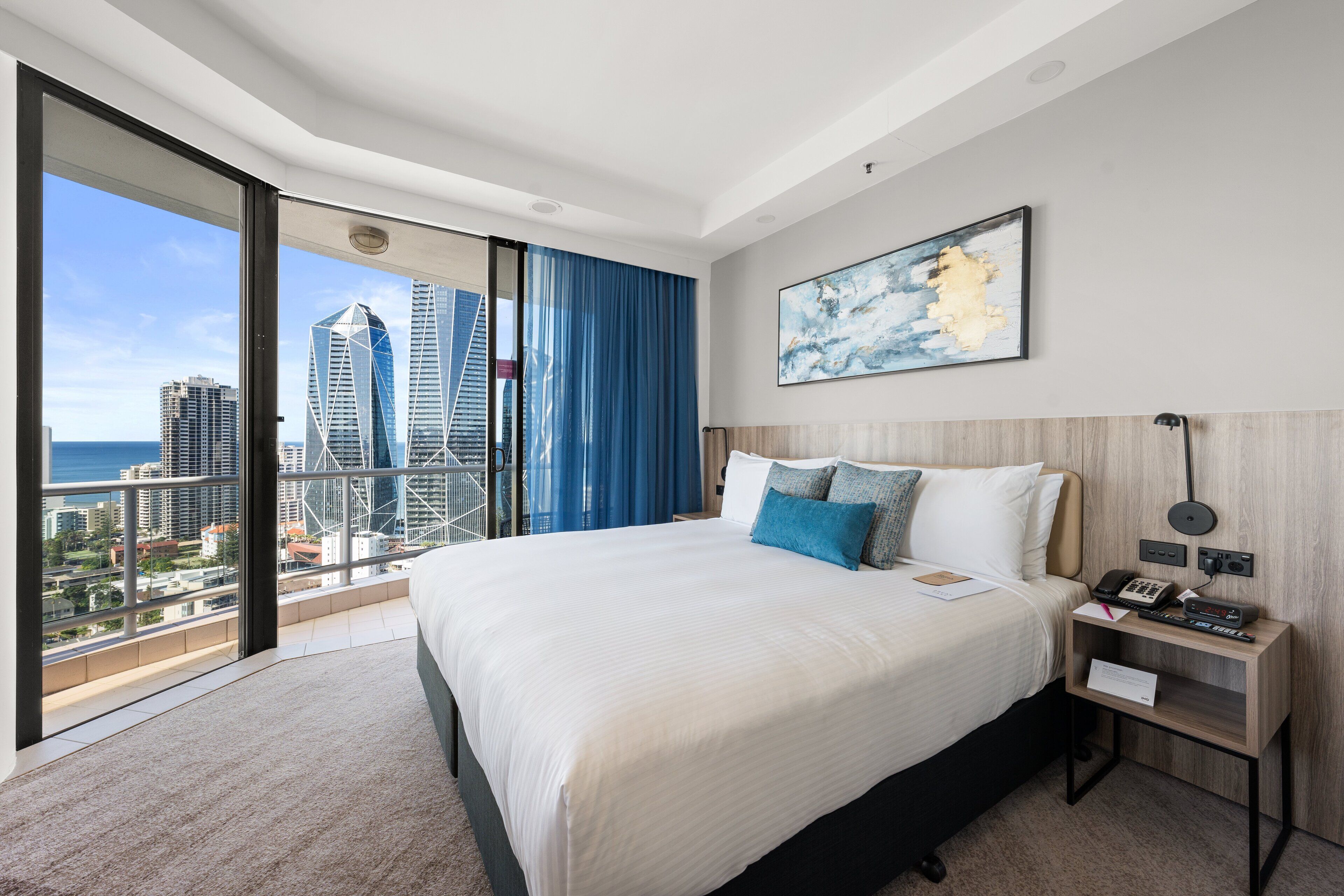 Una habitación de hotel moderna con una cama grande, una mesita de noche con teléfono y control remoto, un cuadro en la pared y un balcón con vista a la ciudad.