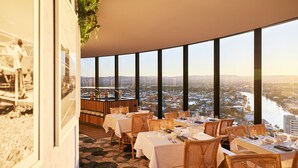 Un restaurante con mesas dispuestas para comer, amplios ventanales que ofrecen vistas a la ciudad y un suelo con diseño de piedra.