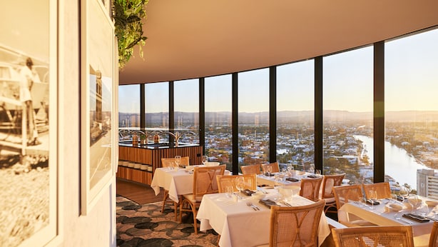 A restaurant with tables set for dining, large windows offering a city view, and a stone-patterned floor.