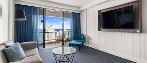 A modern hotel room with a sofa, a round coffee table, a teal armchair, and a large TV mounted on the wall.