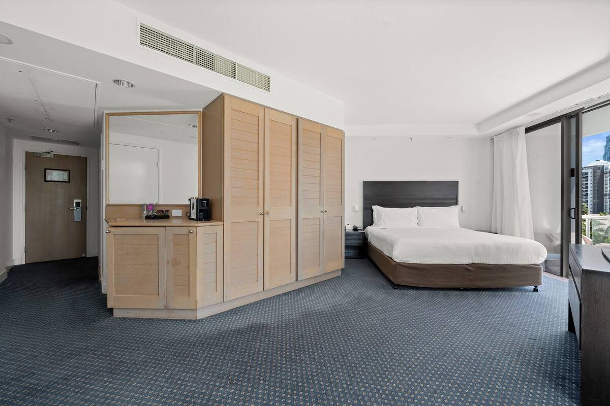A hotel room with a large bed, a wardrobe, a desk with a mirror, and a city view.