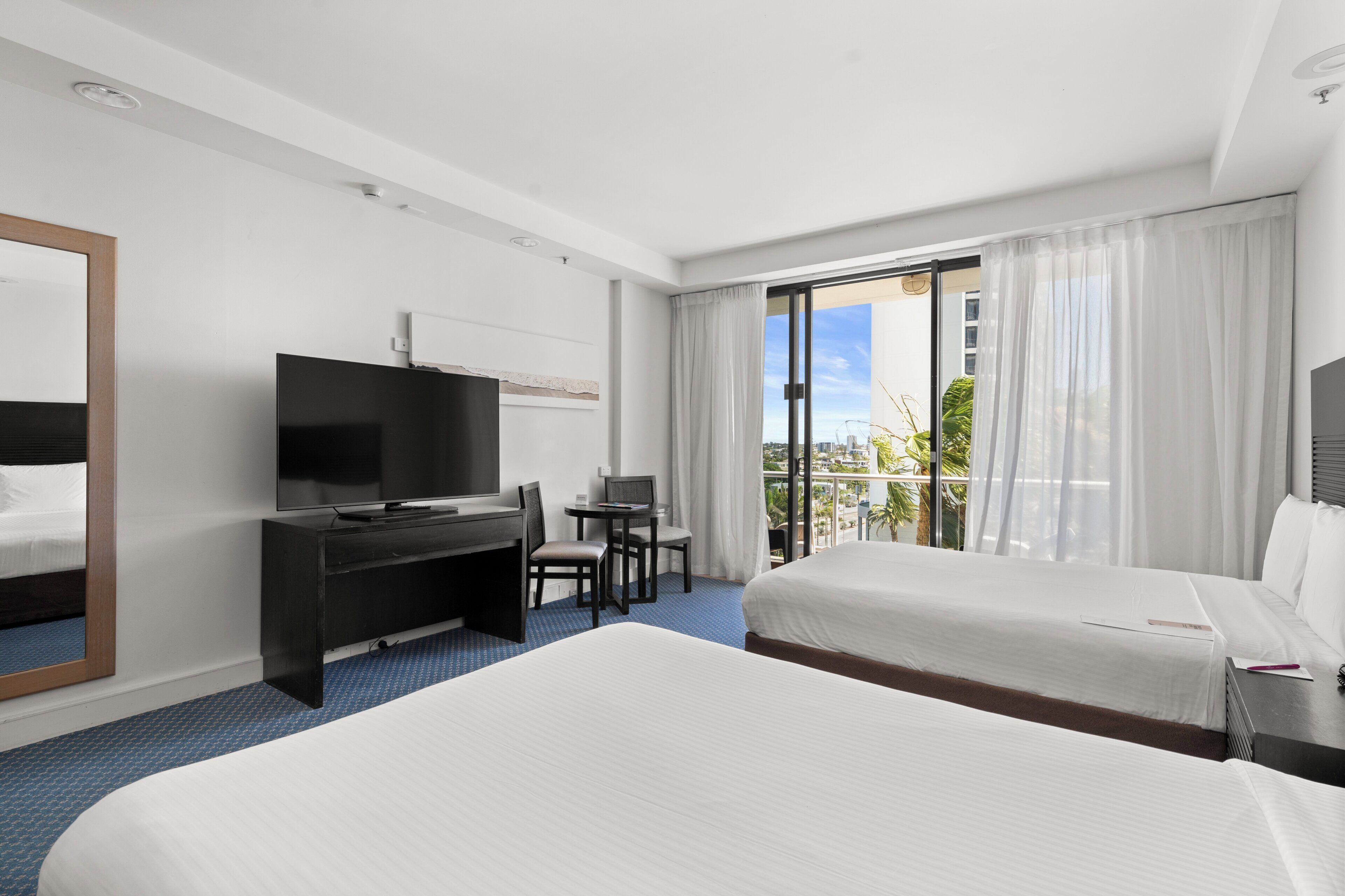 A hotel room with two beds, a desk with a TV, a chair, and a balcony with a view.