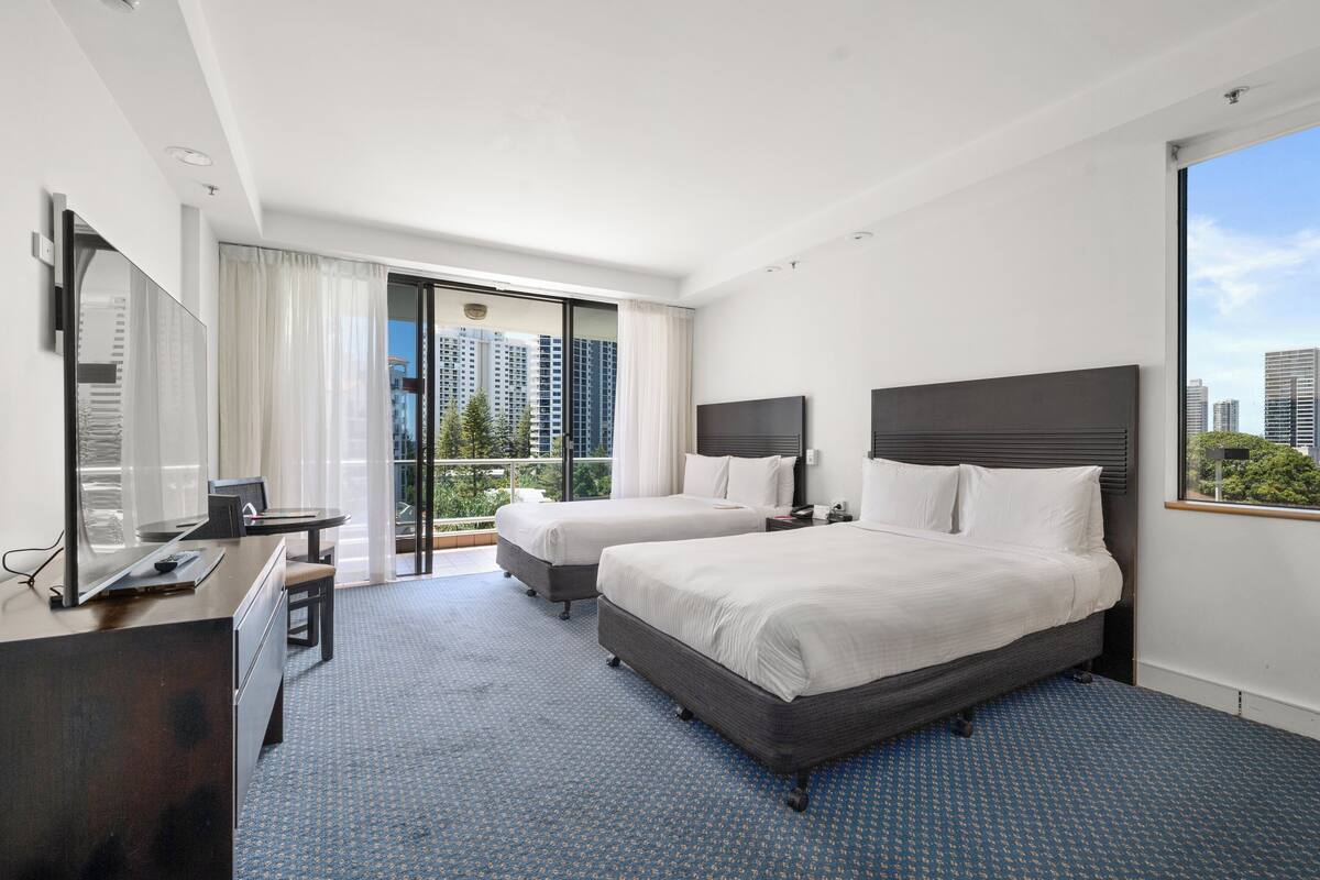 A hotel room with two beds, a desk with a computer, a large window with a city view, and a balcony.