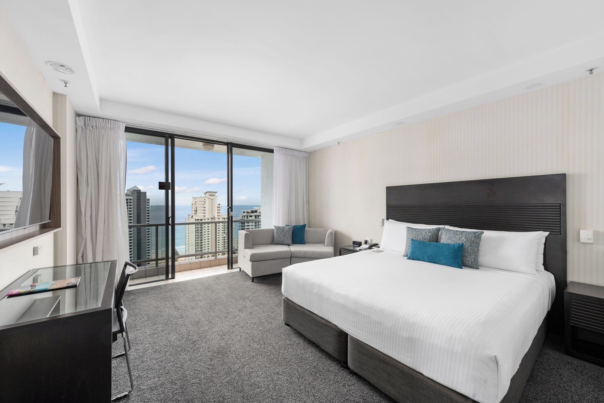A hotel room with a large bed, a desk, a chair, a sofa, and a balcony with a city view.