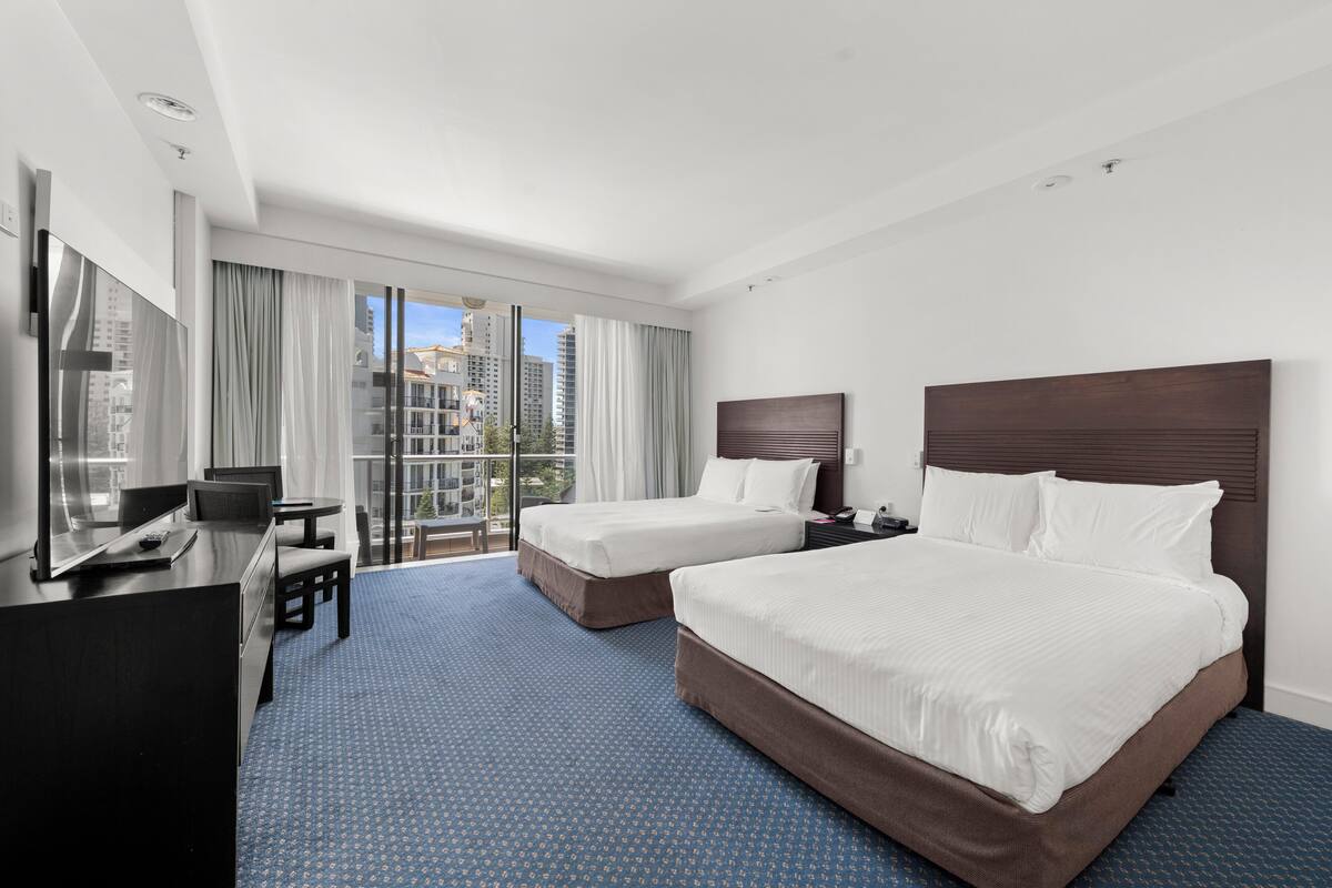 A hotel room with two beds, a desk, and a large window with curtains.