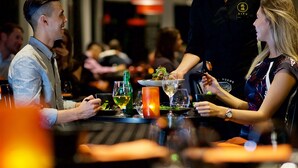 A restaurant setting with diners at tables, a waiter serving food, and a candle on the table.