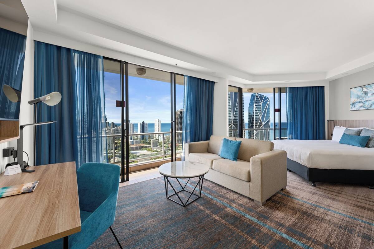 A modern hotel room with a large bed, a sofa, a round coffee table, and a desk. The room has large windows with a city view.