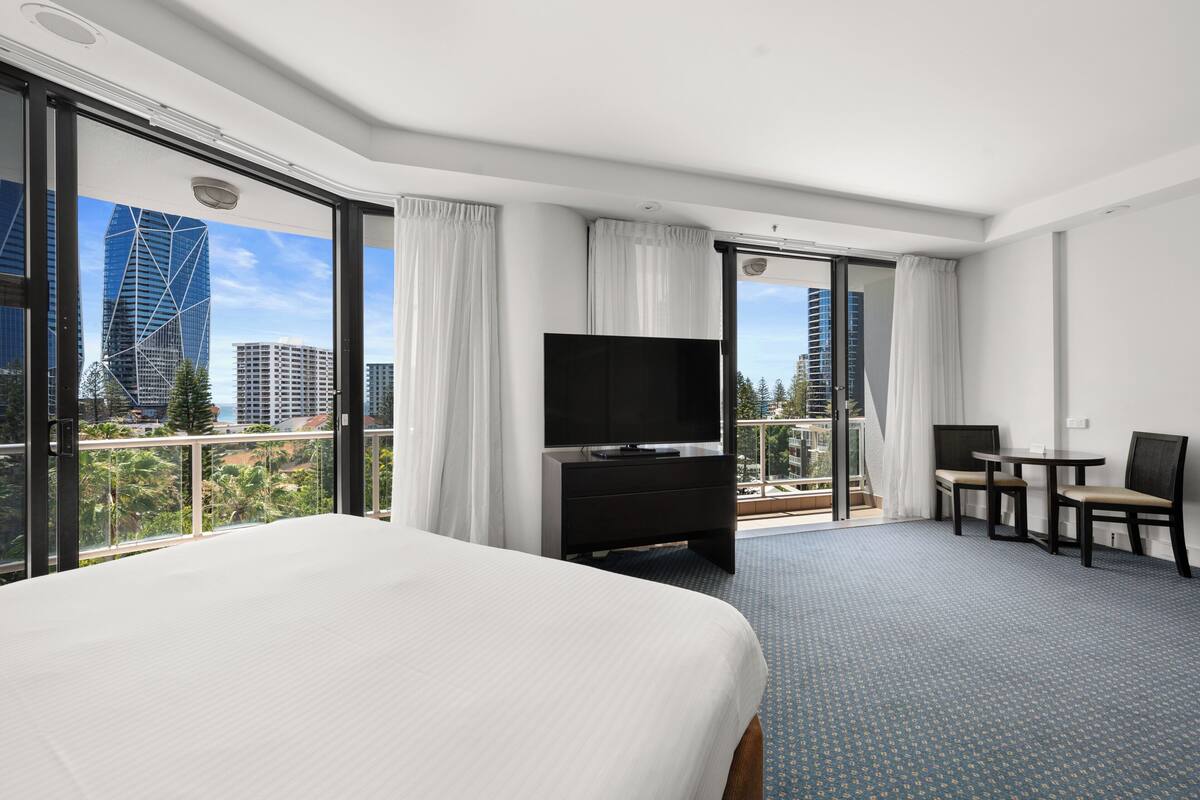 A hotel room with a large bed, a flat-screen TV, a small table with two chairs, and a balcony with a city view.