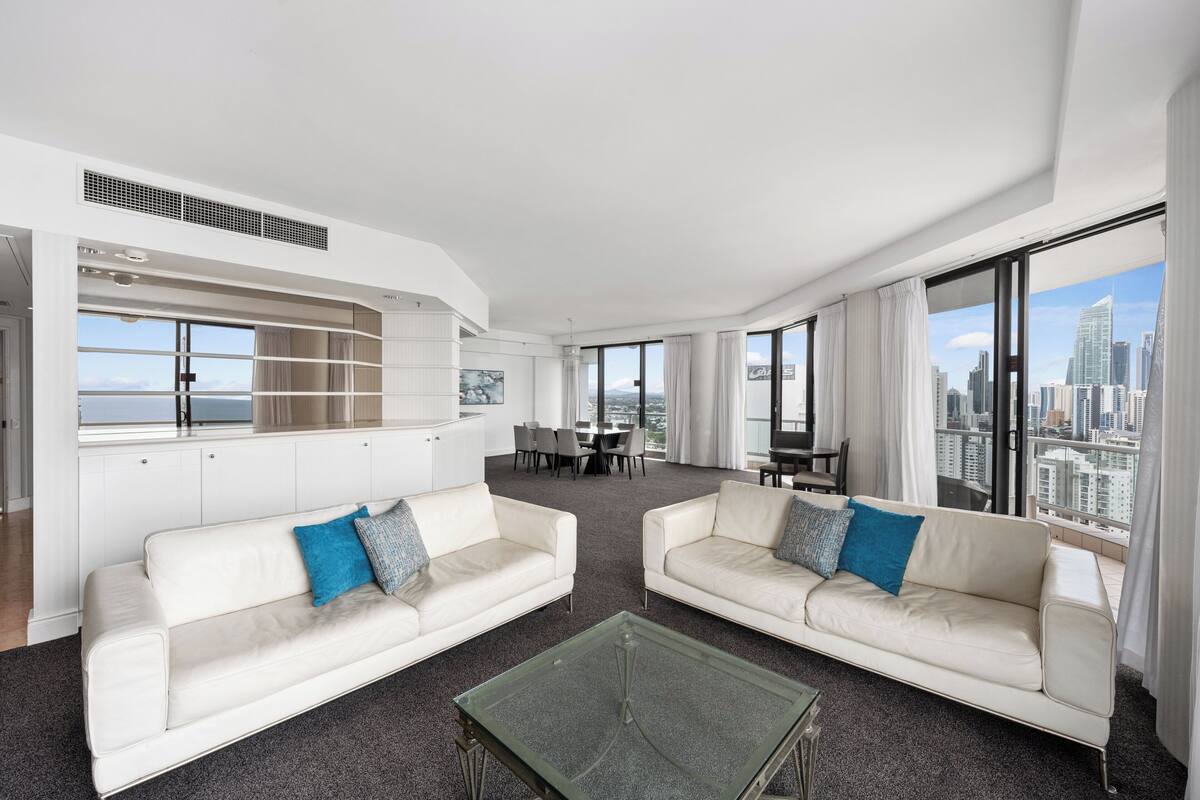 A modern hotel room with a city view, two white sofas with blue cushions, a glass coffee table, and a dining area with a view.