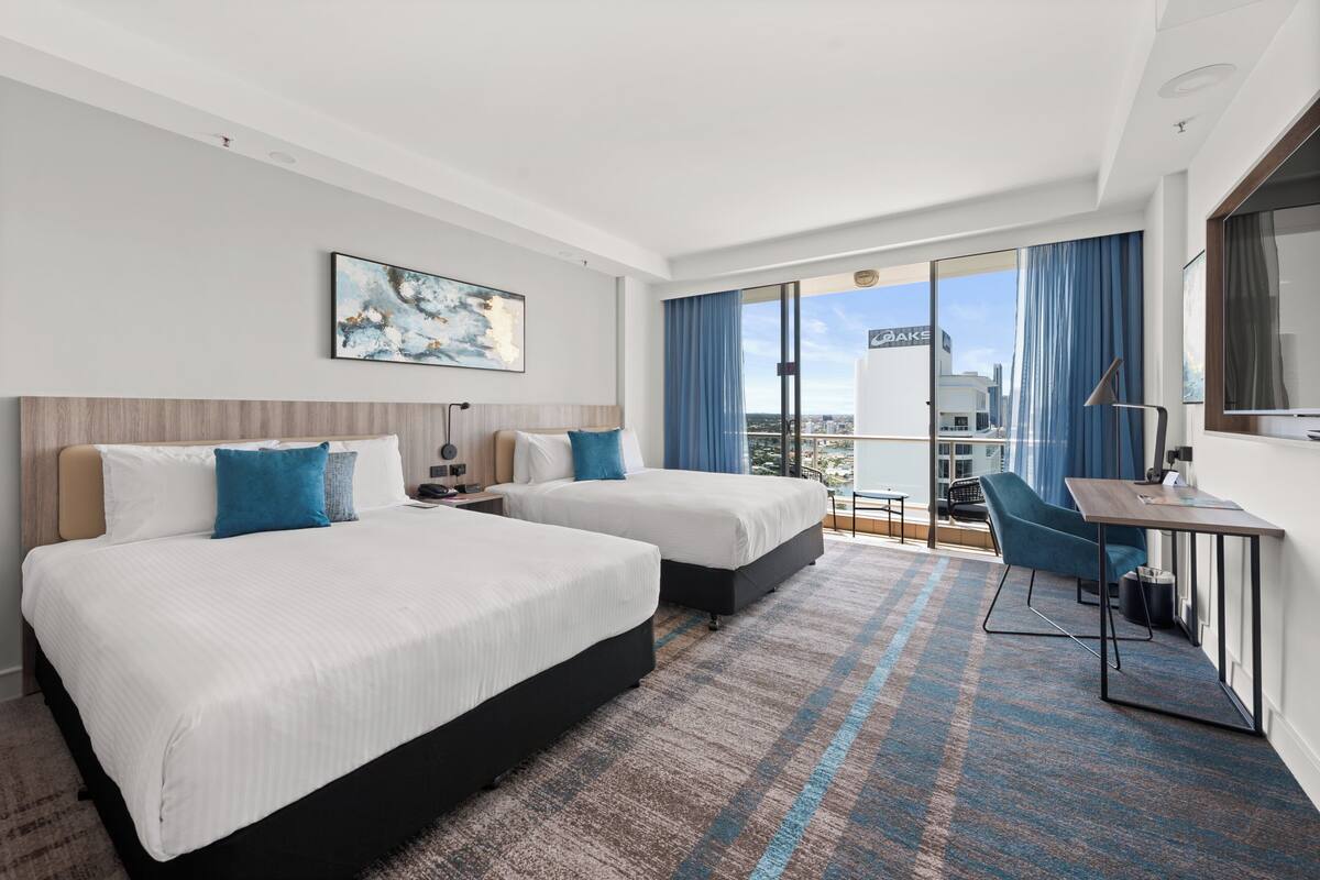 A hotel room with two beds, a desk, a chair, and a large window with a view of a cityscape.