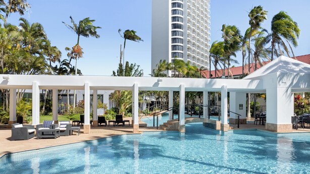 A pool area with lounge chairs and a covered seating area, surrounded by palm trees and a tall building in the background.