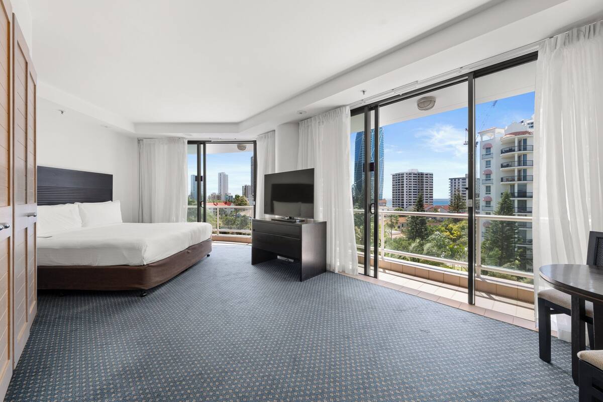 A hotel room with a large bed, a TV, a balcony with a city view, and a small table.