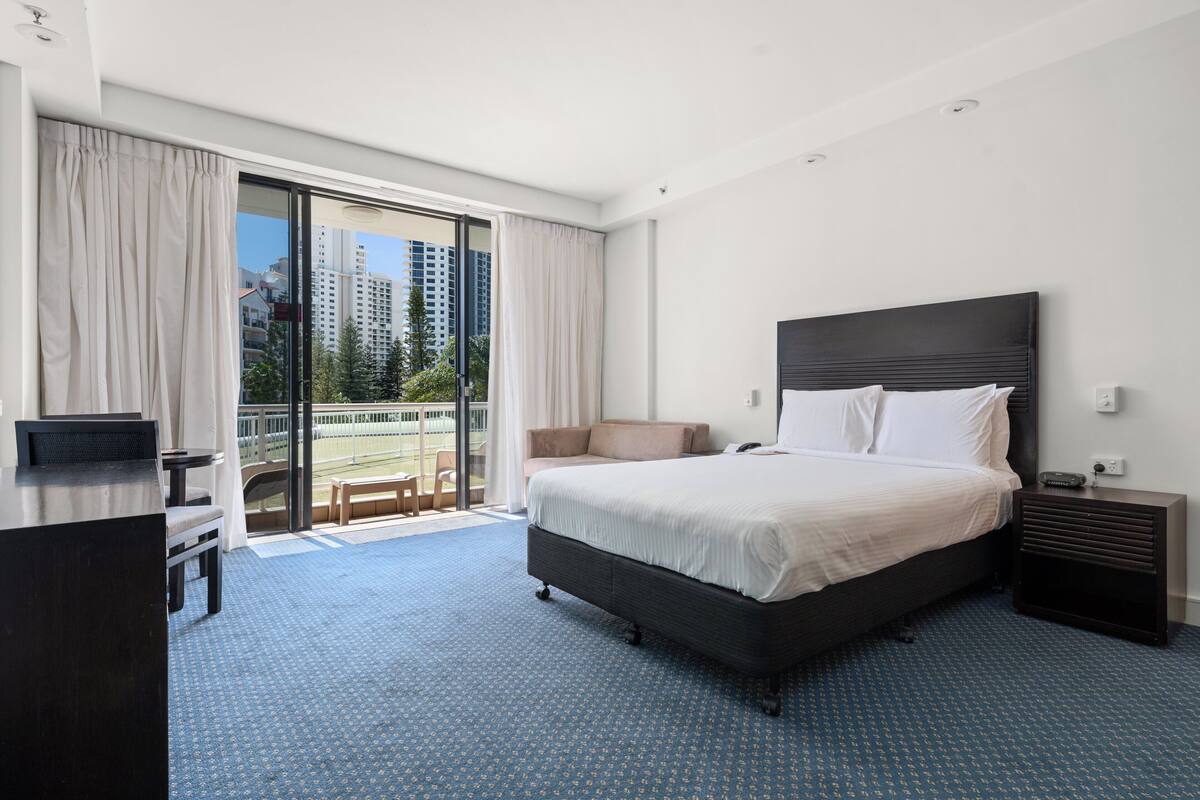 A hotel room with a large bed, a desk, a chair, and a balcony with a view of tall buildings.