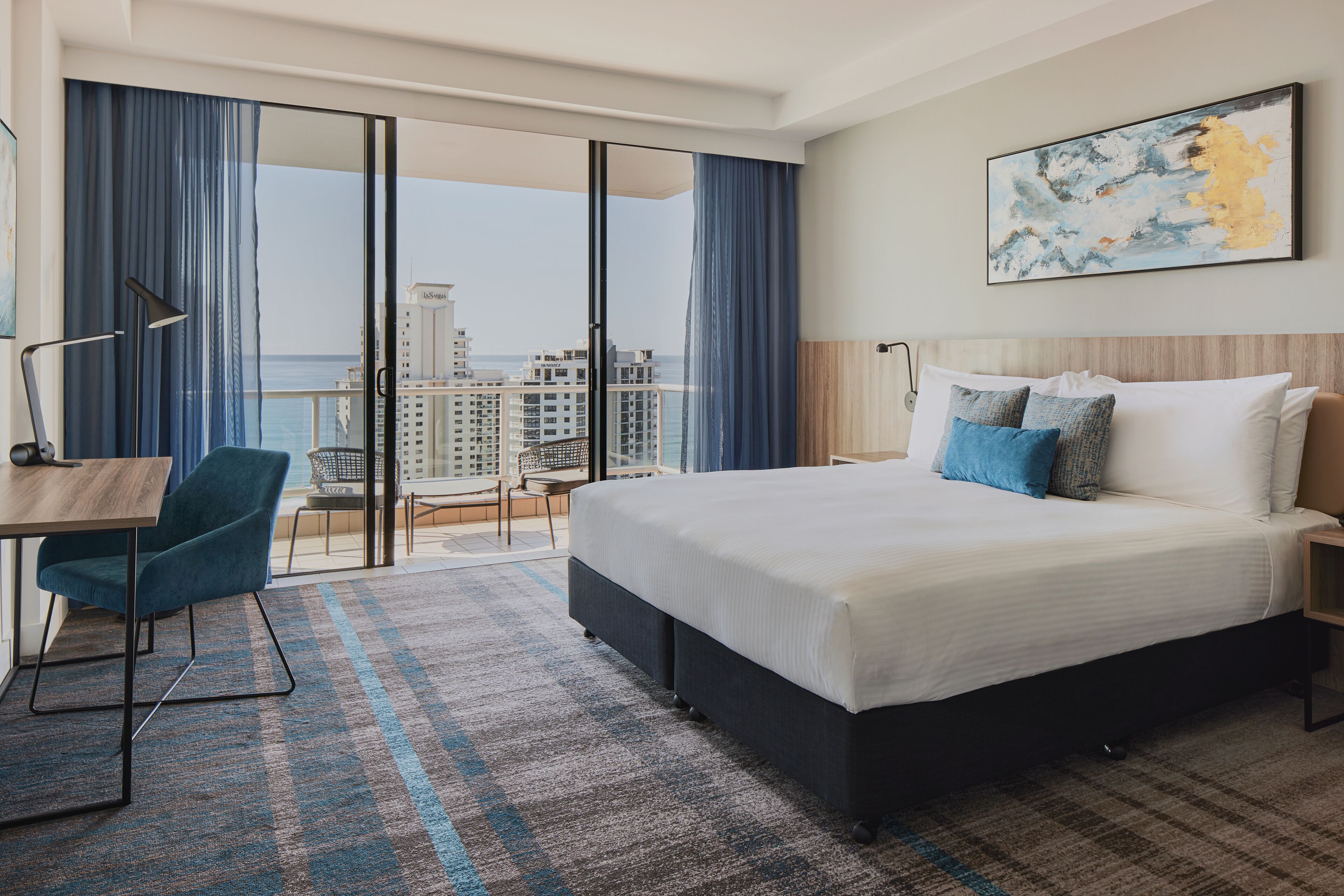 Une chambre d’hôtel avec un grand lit, un bureau, une chaise et une vue sur le paysage urbain, visible à travers une porte vitrée.
