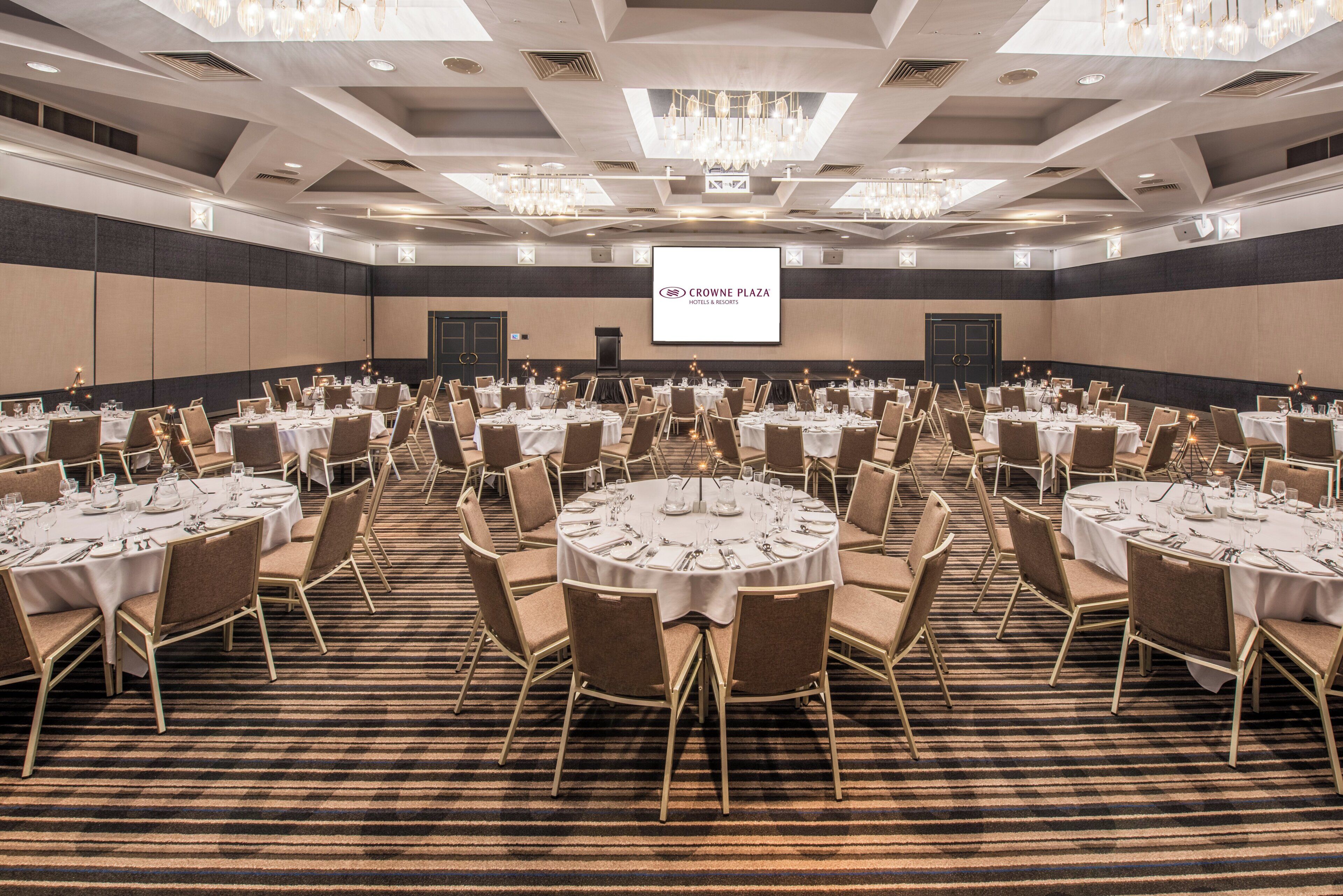 A conference room with round tables, chairs, and a large screen displaying 'CROWNE PLAZA HOTELS & RESORTS'.