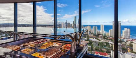 A buffet with a variety of desserts and snacks, a glass railing overlooking a cityscape with buildings and the ocean.