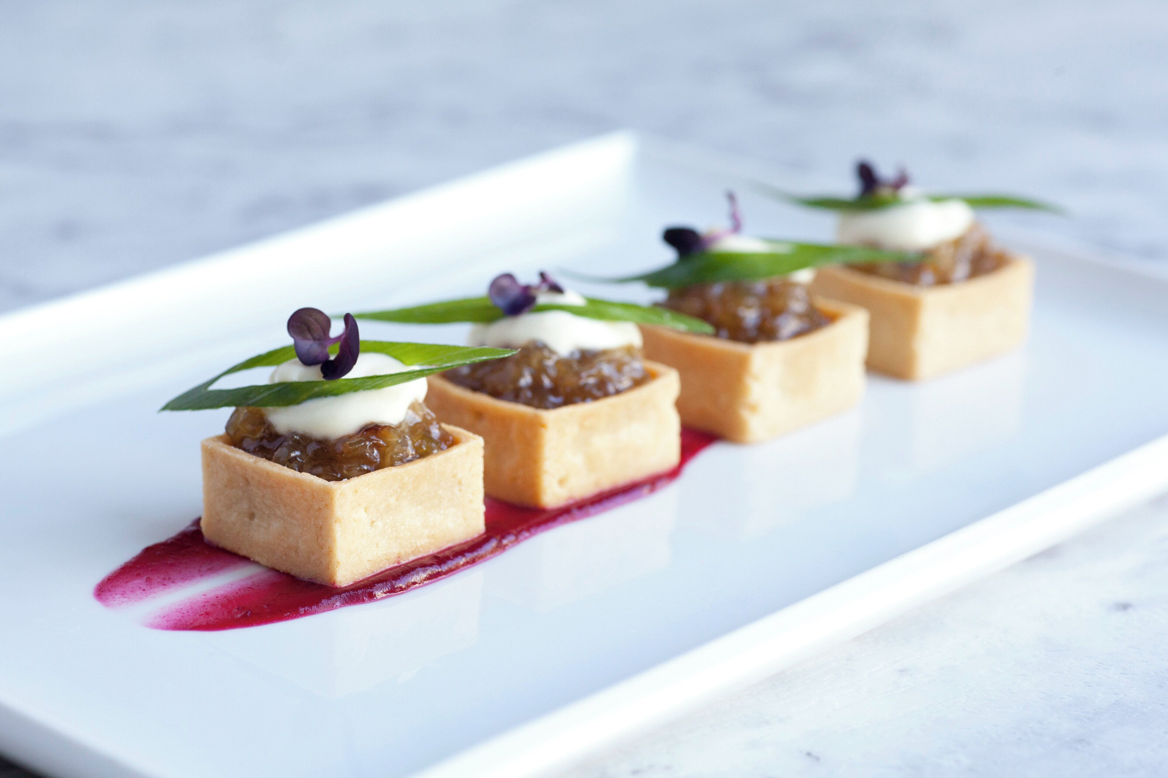 Un plato blanco rectangular con cuatro pastas cuadradas cubiertas de crema y una compota de frutos rojos, decorado con una hoja y una flor morada, sobre una superficie de mármol blanco.