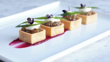 Un plato blanco rectangular con cuatro pastas cuadradas cubiertas de crema y una compota de frutos rojos, decorado con una hoja y una flor morada, sobre una superficie de mármol blanco.
