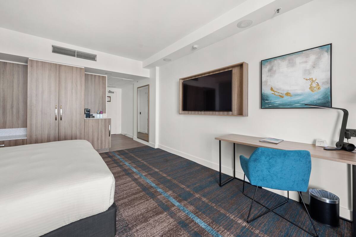 A hotel room with a bed, a desk with a chair, a TV, and a painting on the wall.