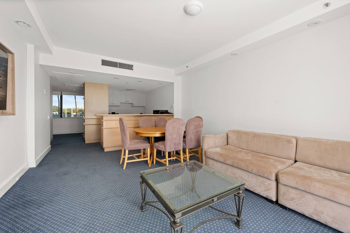 A modern living room with a glass coffee table, beige sofa, and a dining area with wooden furniture.