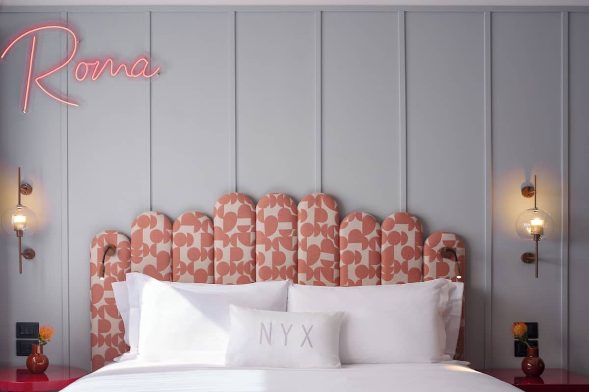 Ein Hotelzimmer mit einem Bett, einem Kopfteil mit Herzmuster, einem leuchtenden Schild mit dem Schriftzug "Roma" und zwei Wandleuchten.