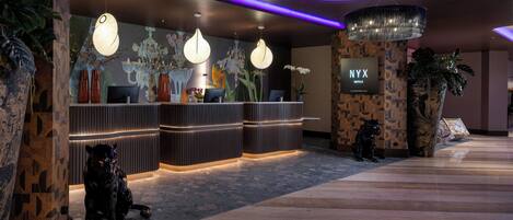 A hotel reception area with a modern design, featuring a reception desk, decorative lighting, and a large chandelier.