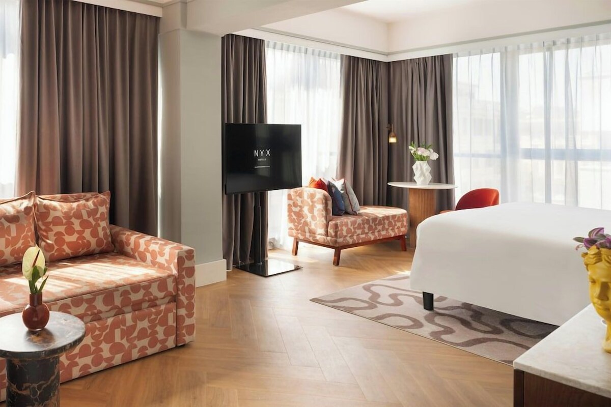Ein Hotelzimmer mit Sofa, Ohrensessel, Bett, Fernseher und einem kleinen Tisch mit einer Vase.