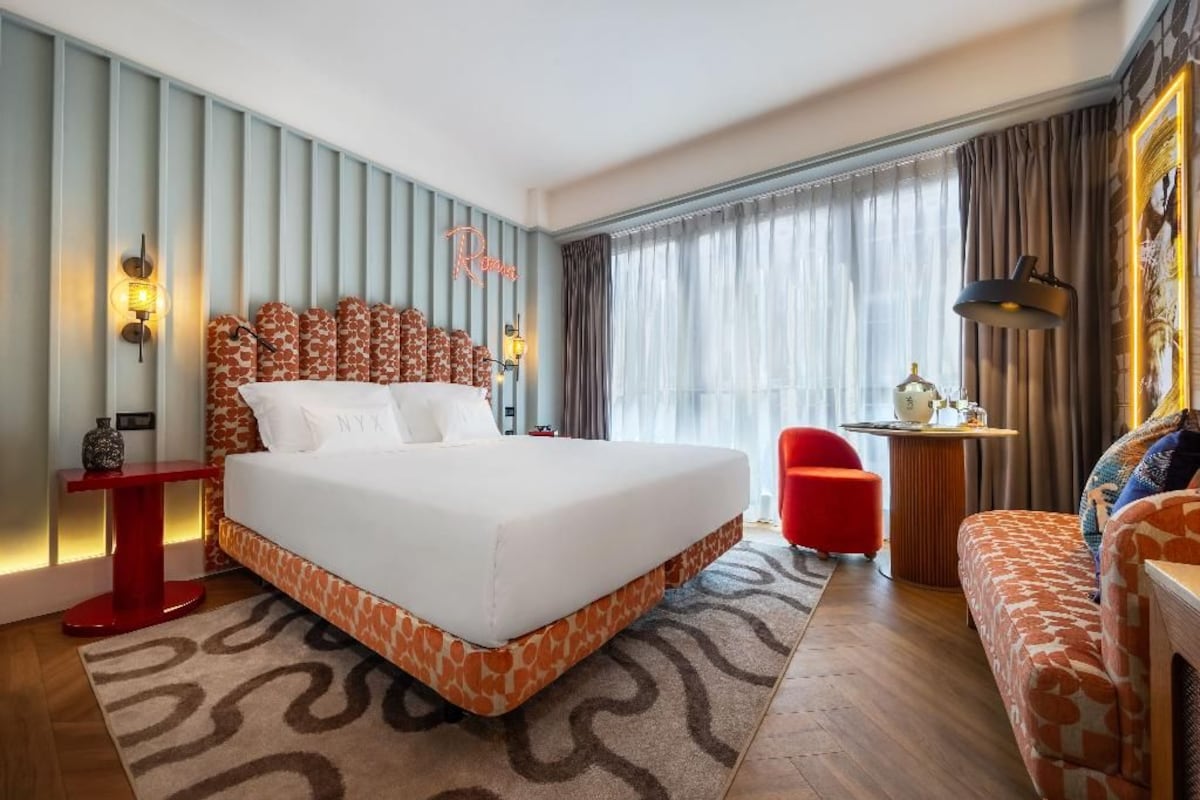 A hotel room with a large bed, a red chair, a sofa, a desk with a lamp, and a window with curtains.