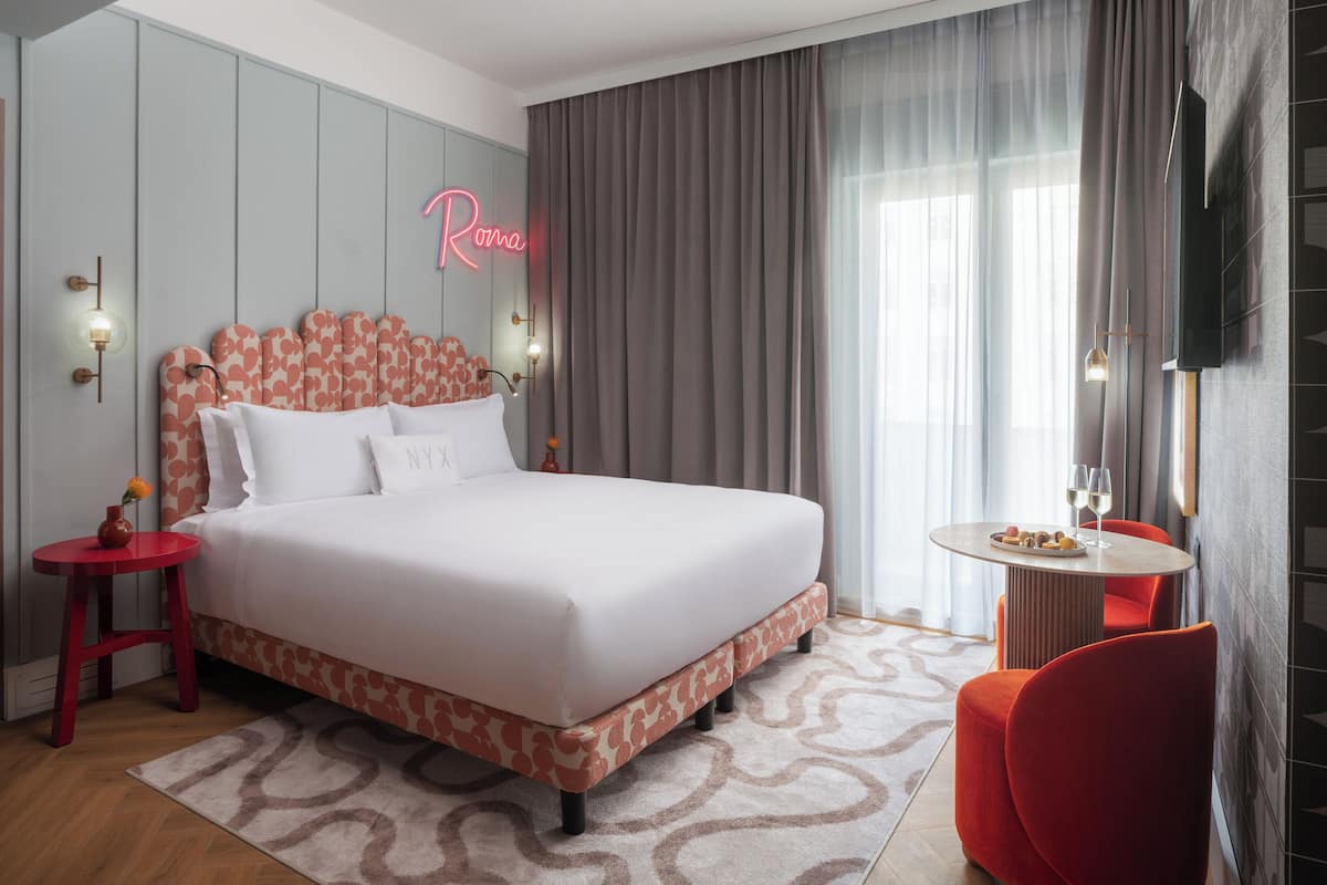 Ein modernes Hotelzimmer mit einem großen Bett, einem roten Sessel, einem kleinen Tisch mit einem Weinglas und einem Neonlicht an der Wand.
