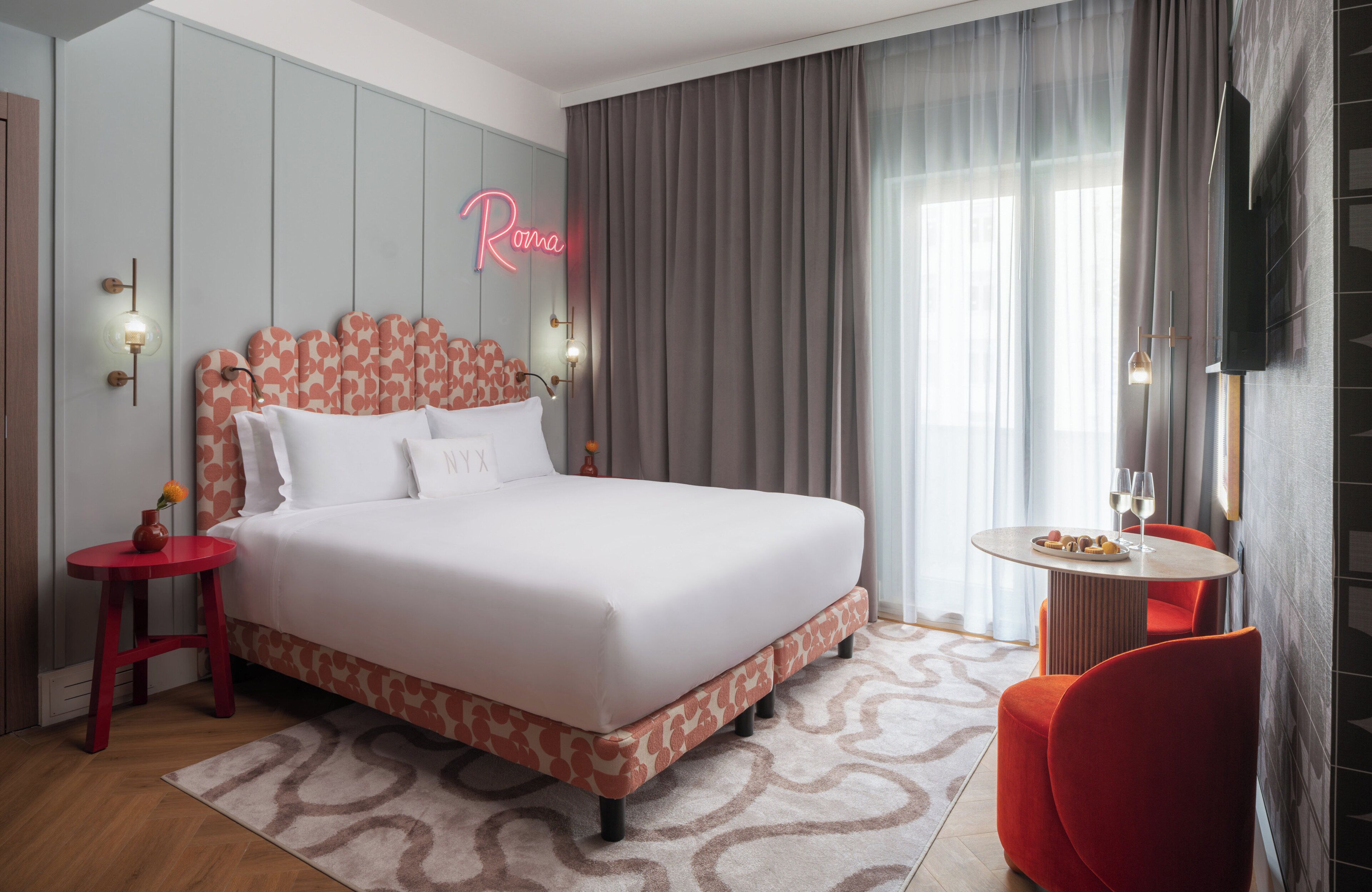 Une chambre d’hôtel moderne avec un grand lit, un fauteuil rouge, une petite table avec un verre de vin et un néon mural.