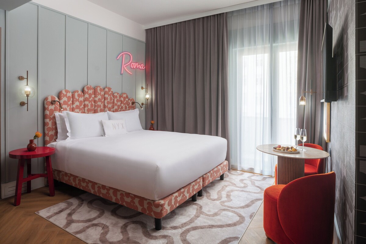 Ein modernes Hotelzimmer mit einem großen Bett, einem roten Sessel, einem kleinen Tisch mit einem Weinglas und einem Neonlicht an der Wand.