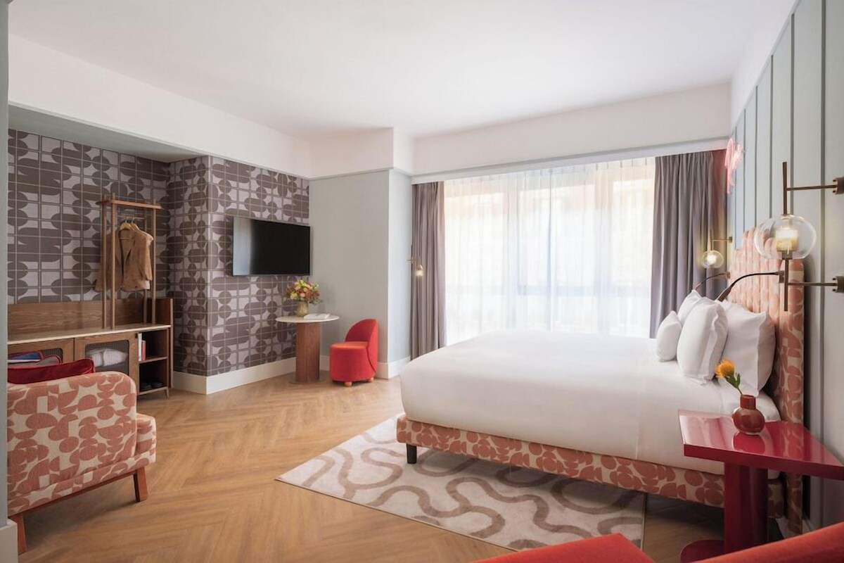 A hotel room with a bed, a TV, a desk, a chair, and a small table with a vase.