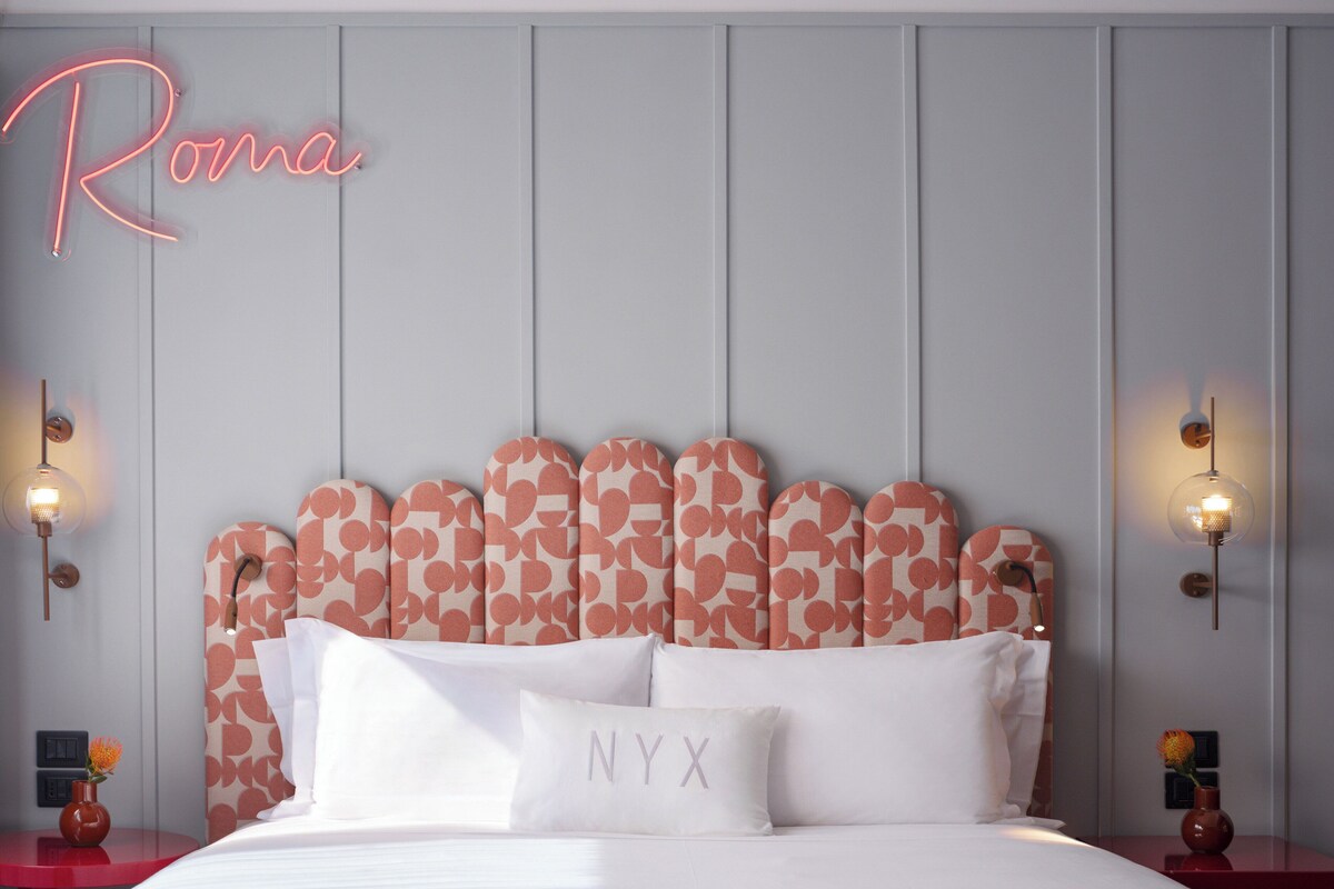 Ein Hotelzimmer mit einem Bett, einem Kopfteil mit Herzmuster, einem leuchtenden Schild mit dem Schriftzug "Roma" und zwei Wandleuchten.