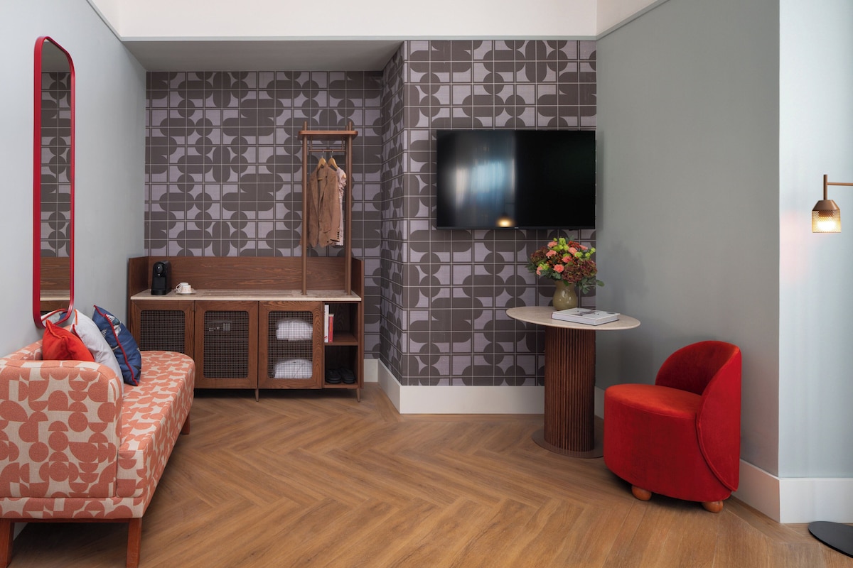 Habitación de hotel moderna con piso de madera en patrón de espiga, un sofá rojo, un sillón rojo, una pequeña mesa redonda con un florero y flores, un televisor montado en la pared, y un diseño de papel tapiz con motivos.