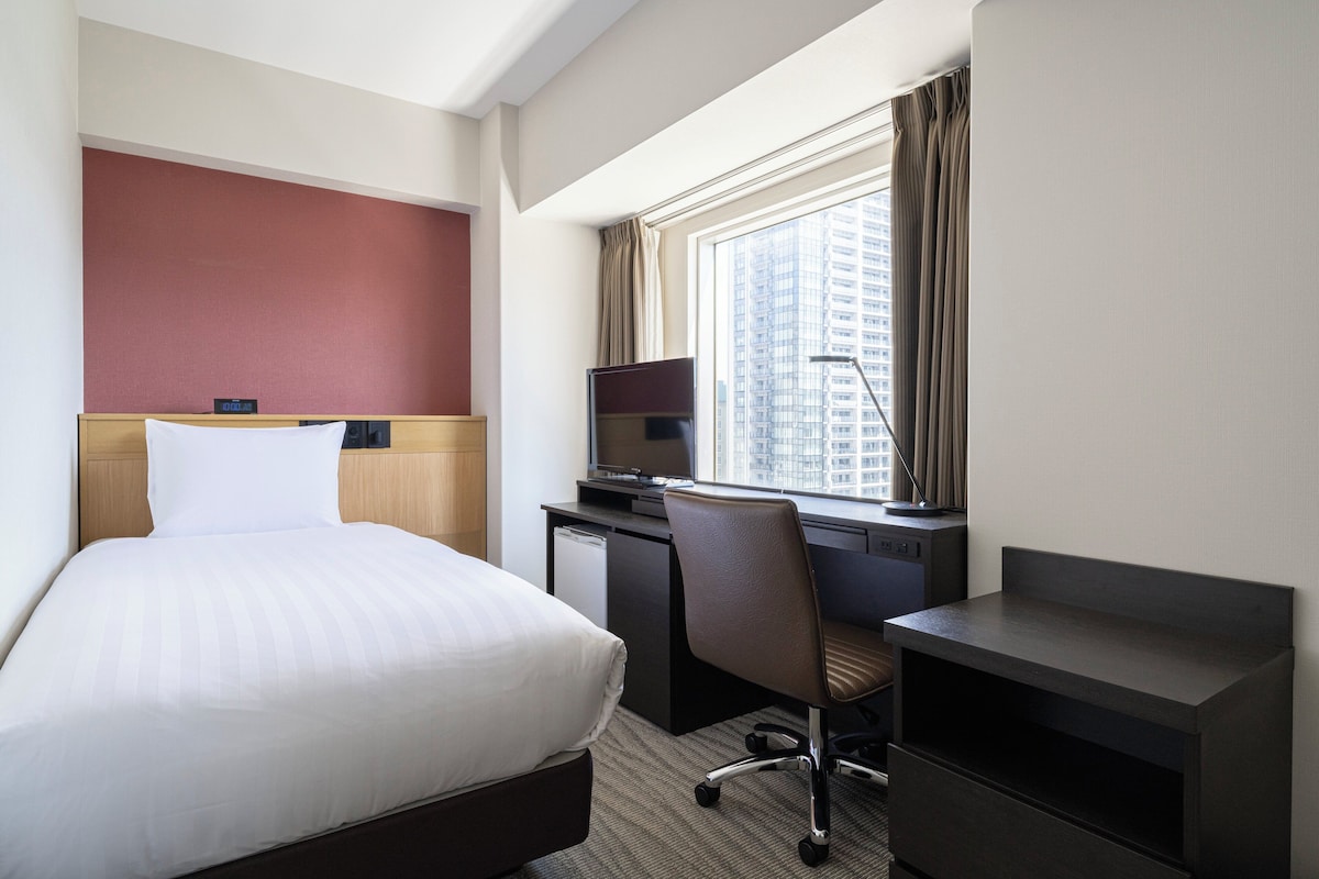 A hotel room with a bed, a desk, a chair, a TV, and a window with a city view.